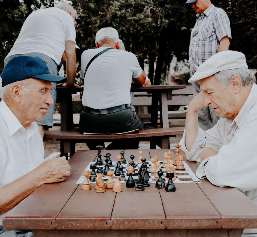 Två äldre herrar spelar schack mot varandra utomhus i en parkmiljö. De sitter vid ett bord i trä