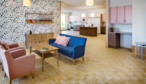 Allmänt utrymme inne på vård- och omsorgsboendet Smedsgården. Rummet är ljust med tapetserade väggar bestående av små svarta löv i svart och ett parkettgolv i trä. Rummet är möblerat med soffa och fåtöljer